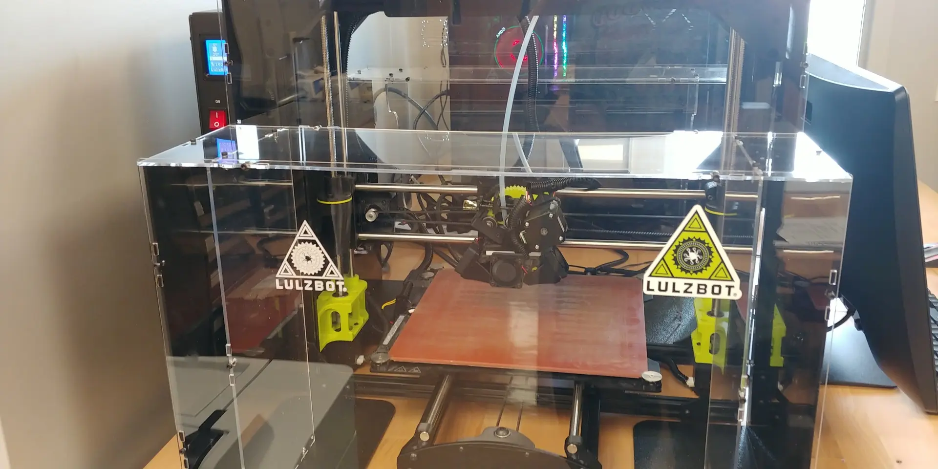 A LulzBot 3-D Printer cased within plexiglass. Shiny steel support bars conjoined to black metal boxes and yellow accessories hover a red print plate.