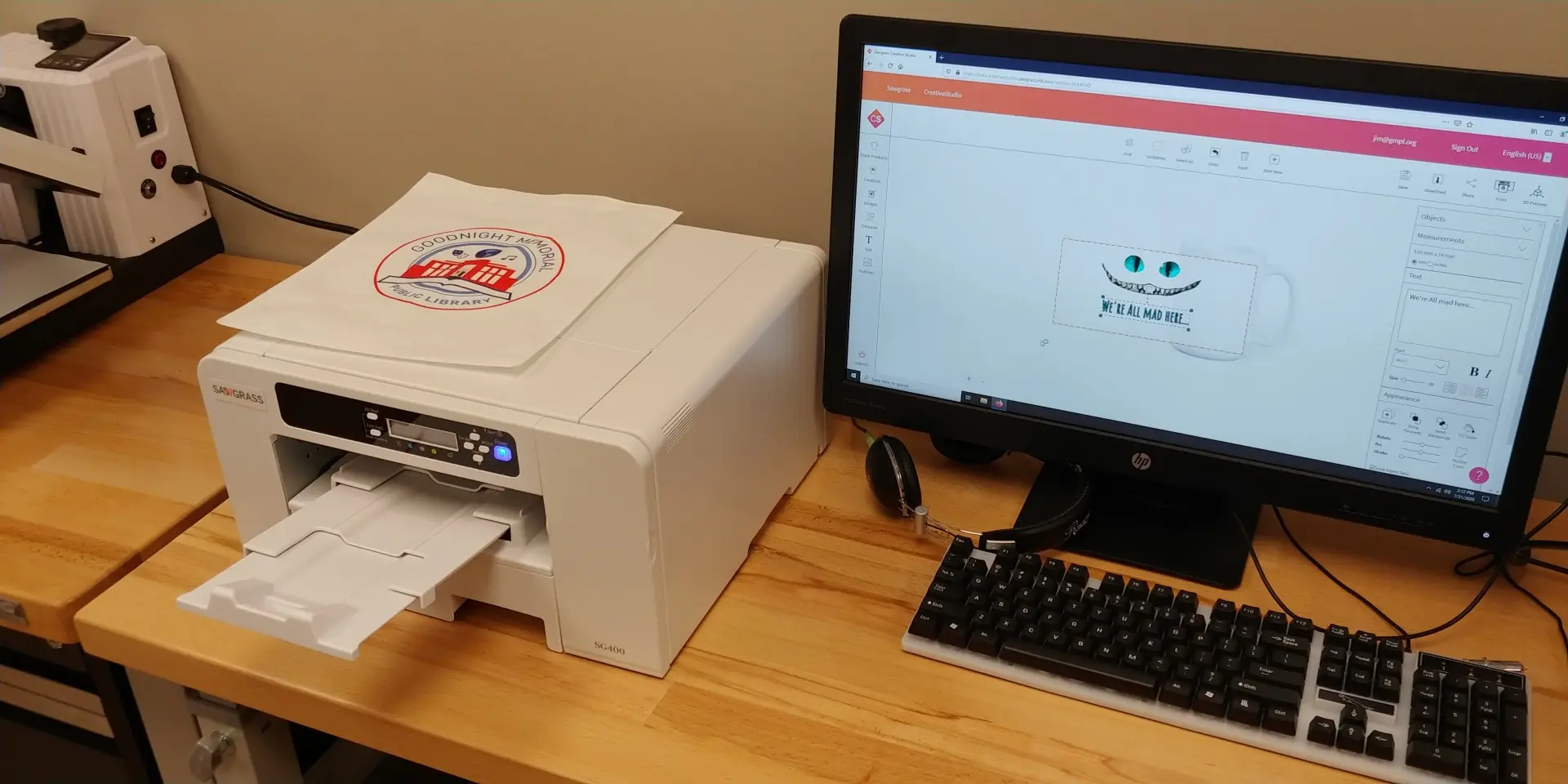 A Beige Dye Sublimation Printer aside a desktop monitor displaying a Cheshire Cat smile on a mug.